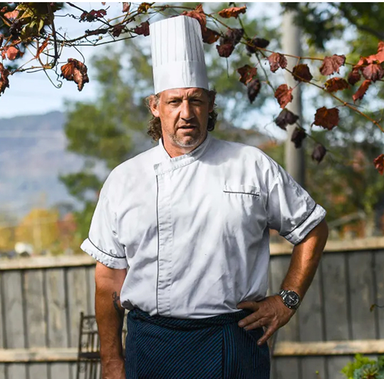 Picture of chef outside wearing a classic chef outfit with a tree in the background with one hand on his hip.